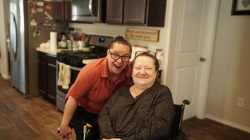 Claudia Verastegui and her mother Norma Verastegui pose together for a photo. Claudia Verastegui slightly bends over/​leans closer to her mom, while Norma Verastegui sits in her wheelchair. Claudia Verastegui makes a funny face, while Norma Verastegui just smiles. – Bild: Warner Bros. Discovery, Inc. or its subsidiaries and affiliates. All rights reserved.