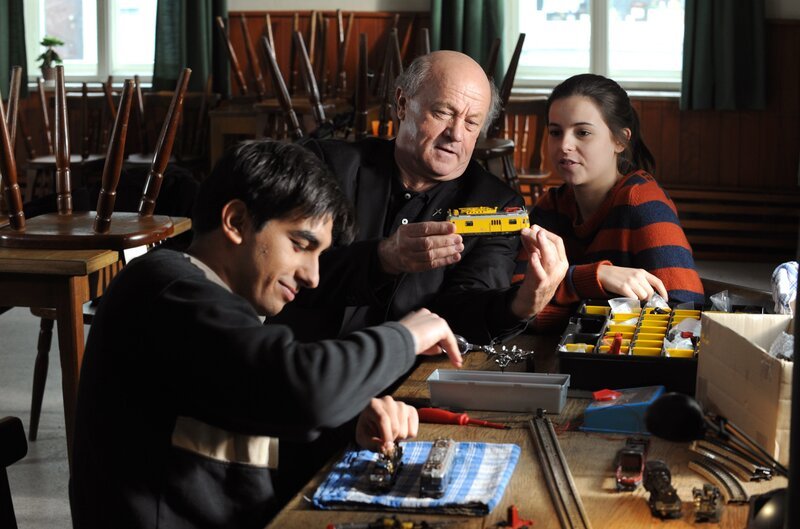 Amir tut die Ablenkung gut. Begeistert nimmt er die Lok unter die Lupe. Von links: Amir (Zejhun Demirov), Pfarrer Kurz (Hans Stadlbauer) und Charlie Keller (Marie-Claire Schuller). – Bild: BR/​Marco Orlando Pichler