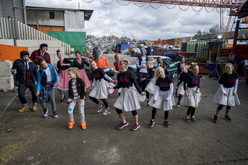 Mit einer fetzigen Rock’n’Roll Nummer sind die beiden Männer abgelenkt und tanzen fröhlich mit Singa, herrH, dem TanzTapir und den TanzAlarm-Kids auf dem Schrottplatz. – Bild: KiKA/​MingaMedia/​Holger Kast
