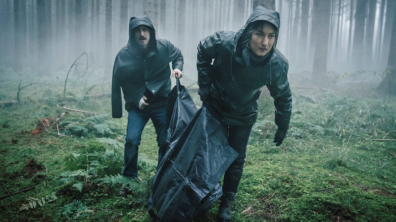 Von links: Ferdinand Zankl (Michael Ostrowski) und Frederike Bader (Marie Leuenberger) tragen Ahmeds Leichensack weg. – Bild: BR/​ARD Degeto/​Hager Moss Film GmbH/​Hendrik Heiden