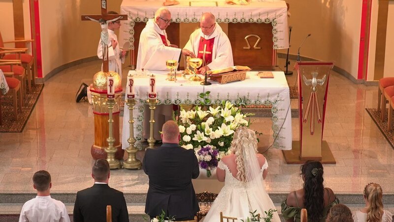 Photograph from the church. The priests are standing behind the altar, the bridal couple is in front of them. The wedding guests are kneeling on the church benches. – Bild: Warner Bros. Discovery, Inc. or its subsidiaries and affiliates