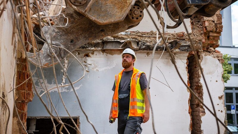 Ein Arbeiter beobachtet, wie ein Bagger Teile des Gebäudes abträgt. – Bild: Warner Bros. Discovery
