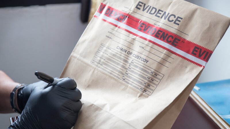 Forensic’s hand in black glove writing on evidence bag and seal by red tape in crime scene investigation – Bild: Discovery/​Prathaan