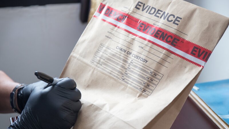 Forensic’s hand in black glove writing on evidence bag and seal by red tape in crime scene investigation – Bild: Discovery/​Prathaan