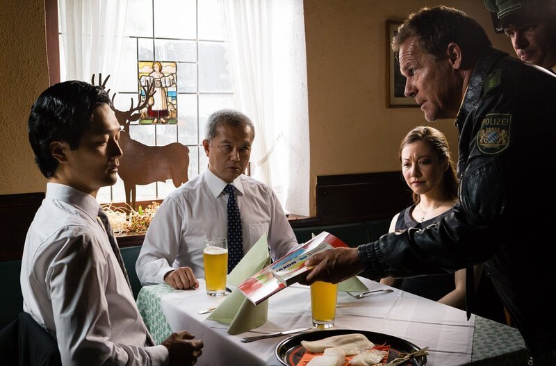 Hubert (Christian Tramitz, 2. von rechts) knallt Momofuku Kasai (Yusuke Yamasaki, links) tiefgefrorenen Fisch auf den Tisch. Er und Staller (Helmfried von Lüttichau, rechts) vermuten, dass der Assistent des Bürgermeisters Takeschi Oshima (Gen Seto, 2. von links) etwas mit dem Verschwinden des wertvollen Kois zu tun hat (mit Nina Fog, 3. von rechts). – Bild: ARD/​TMG/​Arvid Uhlig