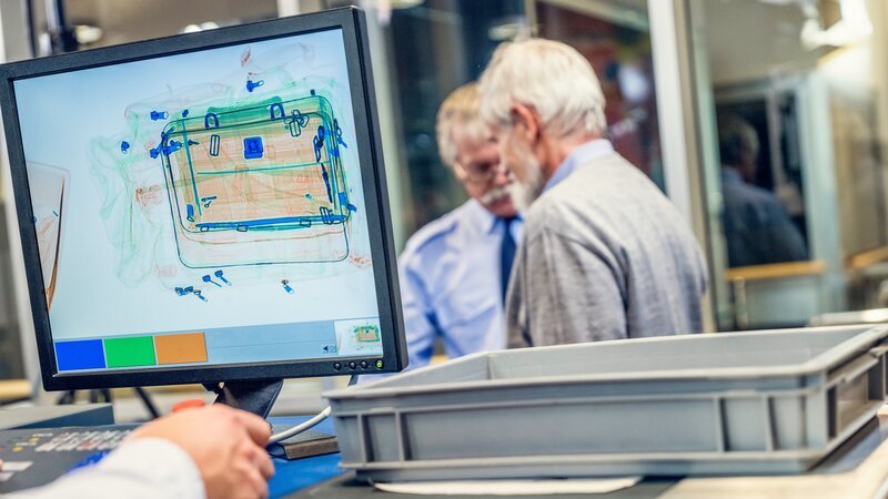 In foreground is computer monitor with luggage image, security guard and senior man in background. – Bild: Model and Property Released (MR&PR)