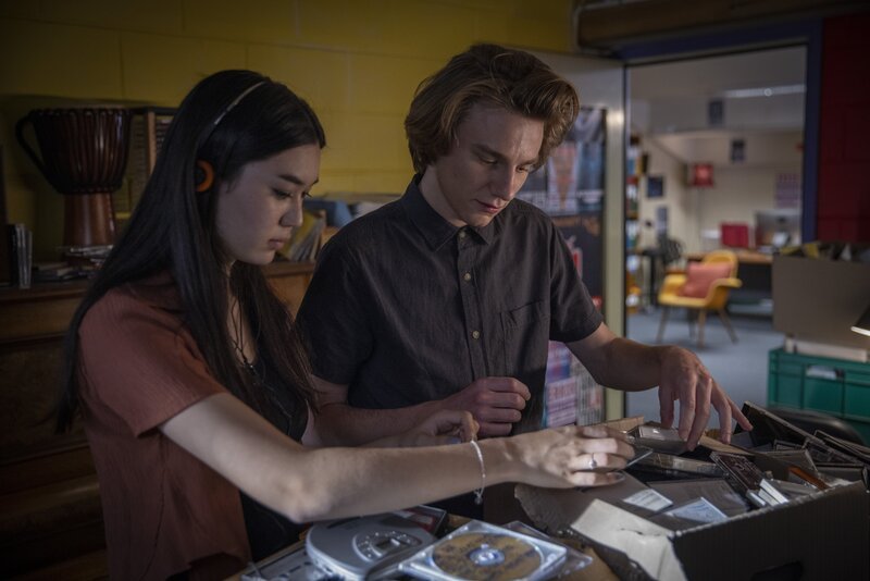 Chester (Henry Strand) und Violetta (Hannah Ogawa) durchstöbern CDs in der Bibliothek. – Bild: BR/​Werner Film Productions/​Ben Saunders