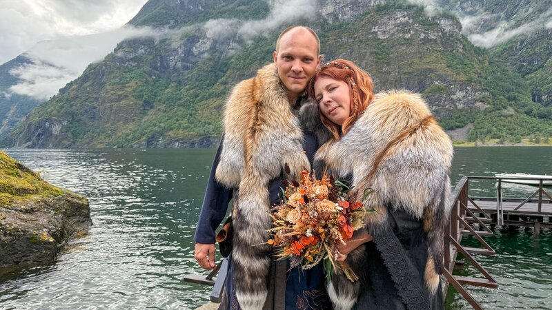 The newlyweds Caroline & Simon are posing in front of the water in their traditional viking wedding gowns. They are cuddled up and looking half smiling into the camera while Caroline is holding her bridal flower bouquet. – Bild: Warner Bros. Discovery, Inc.