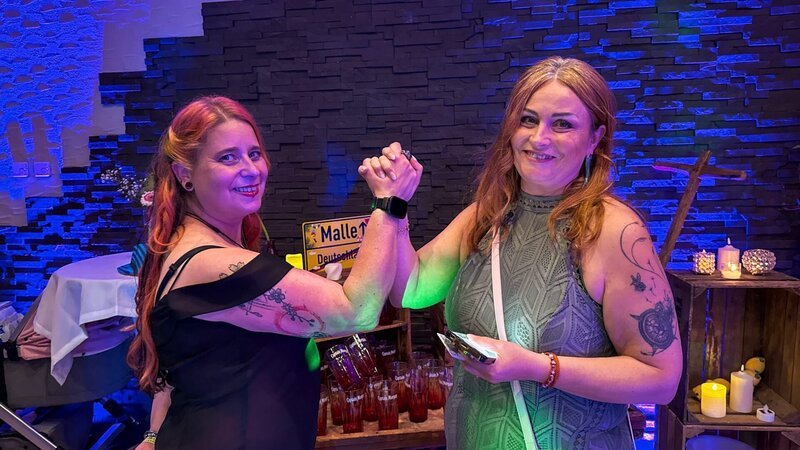 Conversation between wedding planner Christine with her colleague Eva. They stand in front of a movable bar with multiple Captain Morgan glasses. On top is a yellow town sign. – Bild: Warner Bros. Discovery, Inc. or its subsidiaries and affiliates. All rights reserved.