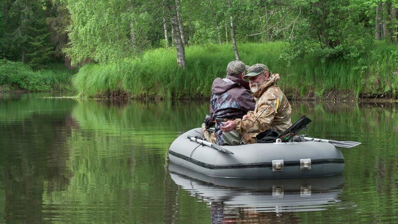 Zwei Personen sitzen in einem Schlauchboot auf einem ruhigen Fluss, umgeben von üppiger grüner Vegetation. – Bild: Waidwerk
