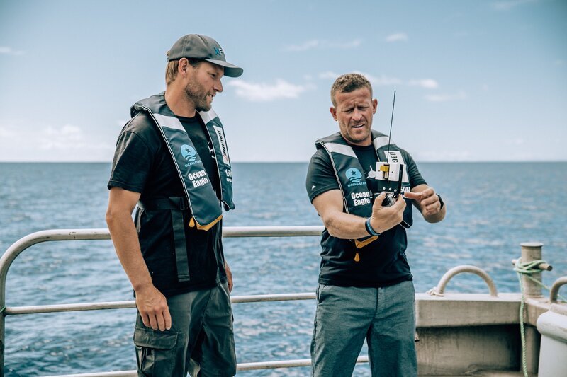 L-R: Riley Elliott und Andy Casagrande diskutieren über die Flossenkamera am Heck des Bootes; Weitwinkelaufnahme. – Bild: Emma Casagrande /​ Discovery Channel /​ Discovery Communications, LLC