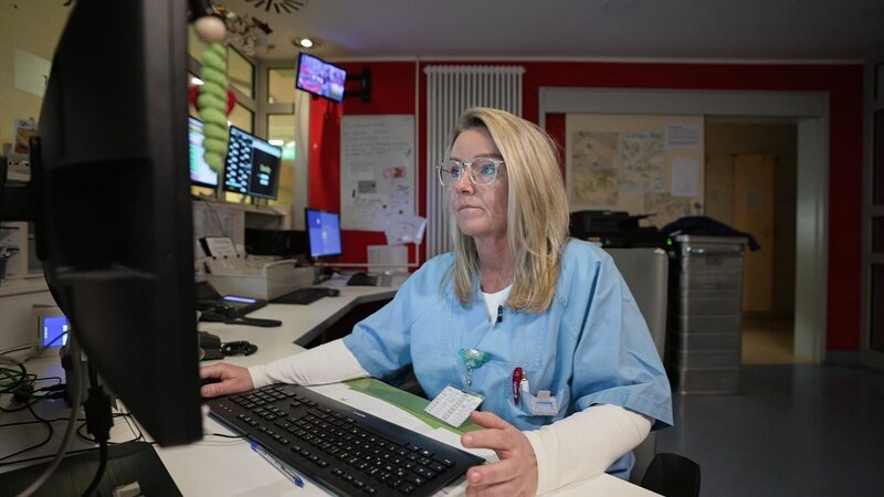 In Magdeburg a middle aged blonde nurse is working at a desk in the staff room. She is wearing glasses and is looking intently at the screen, while her right hand lays on the mouse. Behind her we see a red wall, a big radiatior, a big metallic box among other things. – Bild: Warner Bros. Discovery, Inc. or its subsidiaries and affiliates. All rights reserved.