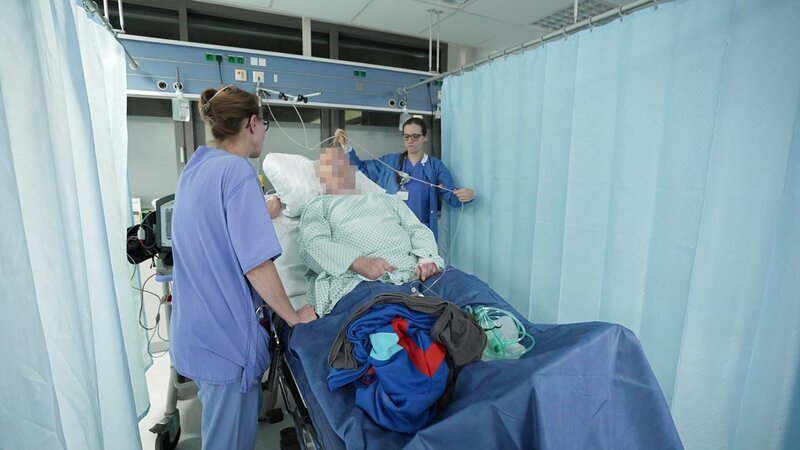 Saarbrücken: Physician Sabine in blue scrubs is standing next to patient in hospital bed, talking to him. Patient has a hospital gown on and his clothes are lying on the bed. On his right side nurse Sabrina is handling his IV. On both side are blue curtains. – Bild: Warner Bros. Discovery, Inc.