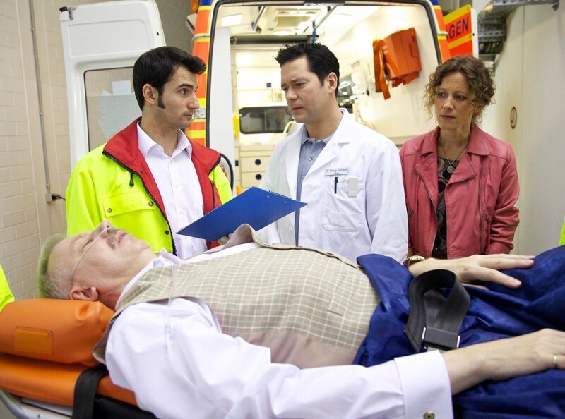 Gero Kramer (Ludger Pistor, liegend) hat gegen die ärztlichen Anweisungen von Dr. Brentano (Thomas Koch, Mitte) die Klinik verlassen. Seine Auszubildende Beate Weiß (Katharina Speiring) hat dem von Zwängen besessenen Gero gesagt, sie hätte seine Wohnungstür nicht abgeschlossen. Gero blieb nichts anderes übrig, als dies selbst zu kontrollieren. Das Ergebnis: Gero ist wieder zusammengebrochen und wird nun erneut vom Notarzt (Roman Petermann, links) eingeliefert. Trotz der Verärgerung über ihren Chef, hat Beate Weiß ein sehr schlechtes Gewissen, weil sie Gero derartig provoziert hat. – Bild: MDR/​Wernicke