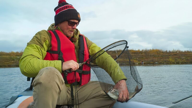 Eine Person im Boot hält ein Netz mit einem gefangenen Fisch auf einem ruhigen Fluss. – Bild: Waidwerk