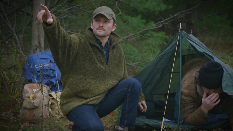 The man pointing in the wilderness, while another person is seen distressed inside a tent. – Bild: Warner Bros. Discovery