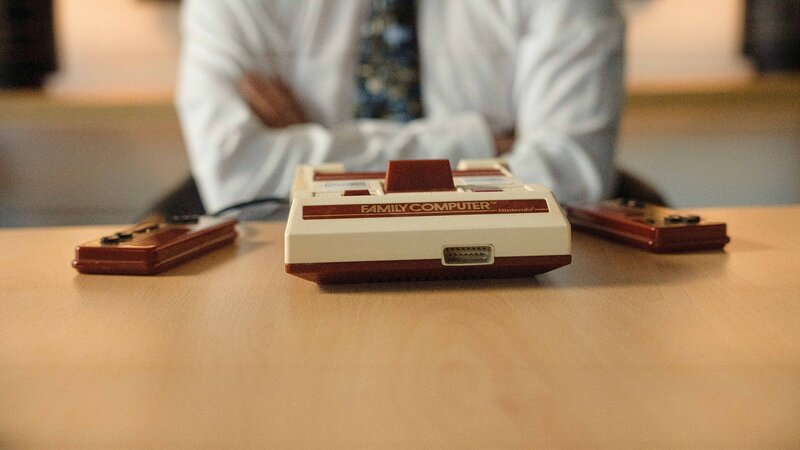 Auf dem Tisch steht eine Retro-Konsole „Family Computer“ mit zwei Controllern, während eine Person im Hintergrund mit verschränkten Armen sitzt. – Bild: aetnsta /​ THE HISTORY CHANNEL /​ A+E Networks