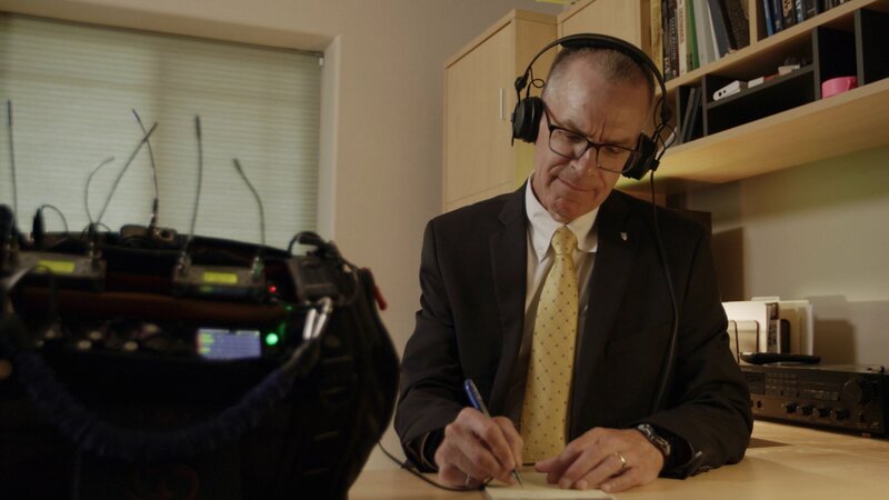 Eine Person im Anzug sitzt am Schreibtisch, trägt Kopfhörer und schreibt auf Papier, während vor ihr Audioausrüstung mit Kabeln steht. – Bild: A+E Global Media /​ THE HISTORY CHANNEL