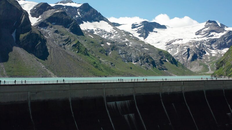 Blick auf eine Staumauer vor einem Bergsee, umgeben von hohen schneebedeckten Gipfeln. – Bild: Kurt Reindl Filmproduktion