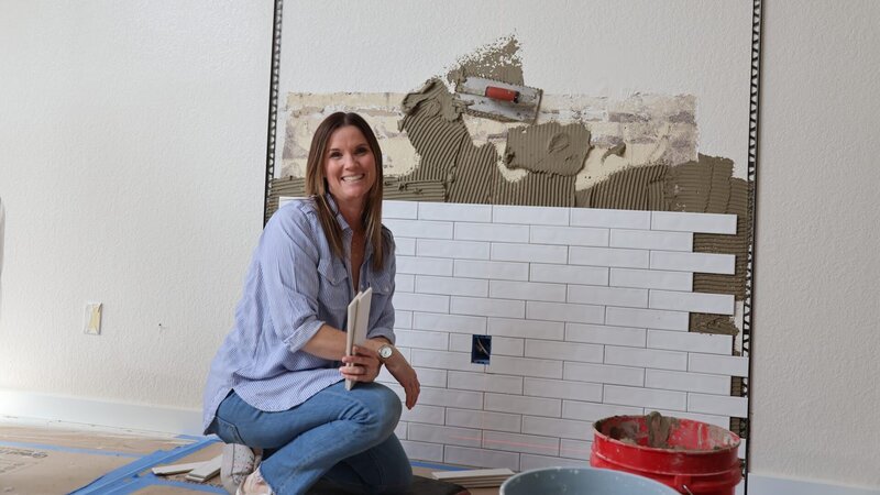 Leslie poses with tile in front of the in-progress fireplace backdrop – Bild: Warner Bros. Discovery, Inc. or its subsidiaries and affiliates. All rights reserved.