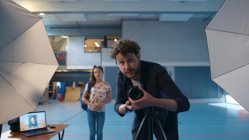Der Schulfotograf (Thomas Höppener) macht ein Foto von Floor (Bobbi Mulder), während Margeet (Romy Voll) im Hintergrund zuschaut. – Bild: NDR/​NL Film