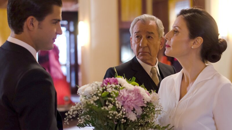 Dona Blanca und Don Emilio (Mitte, José Sacristán) beauftragen den jungen Verkäufer Max (l., Maxi Iglesias), Cristina (r., Aitana Sánchez-Gijón) anlässlich der Rückkehr von ihrer Hochzeitsreise einen Blumenstrauß zu überreichen. – Bild: WDR/​Enrique Baró Ubach/​Beta Film GmbH