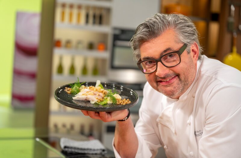 Starkoch Hans Jörg Bachmeier steht in der Studioküche. Er hat einen Teller in der Hand und probiert das Gericht.; Starkoch Hans Jörg Bachmeier hält einen Löffel und ein Tellergericht in der Hand.; Starkoch Hans Jörg Bachmeier präsentiert ein Tellergericht. – Bild: BR/​Ralf Wilschewski