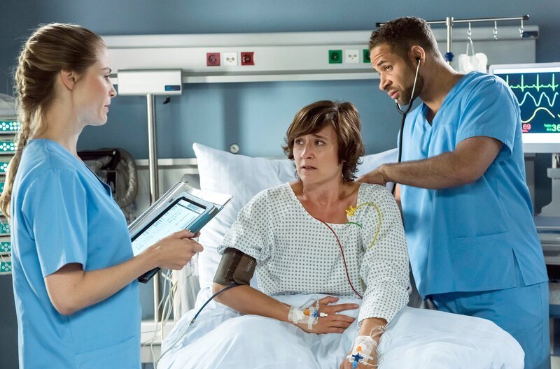 Die Ärzte vermuten eine Vergiftung; Julia (Mirka Pigulla, l.) entnimmt Frau Brücker (Magdalena Helmig, M.) erneut Blut, während Matteo (Mike Adler, r.) die Lungen prüft. – Bild: ARD/​Jens Ulrich Koch