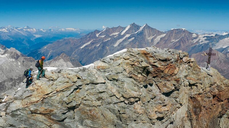 Menschen steigen über einen felsigen Bergrücken mit weitem Blick auf die umliegenden Gipfel. – Bild: Waidwerk