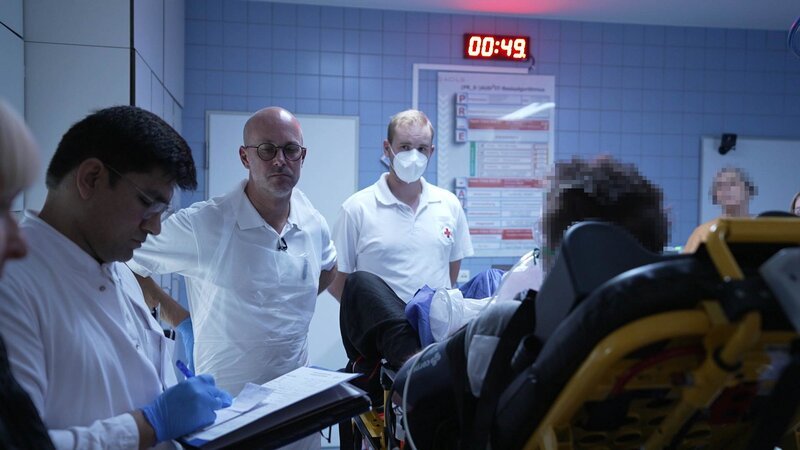 Kaufbeuren: Ein Patient (rückseitig mit verpixeltem Kopf) liegt halb auf einer Bahre im Schockraum; mehrere Mitarbeiter und ein maskierter Rettungsdienstmitarbeiter sind zu sehen; an der Wand ein Digitaltimer. – Bild: Production Company Staff /​ DMAX – Germany /​ Warner Bros. Discovery, Inc.