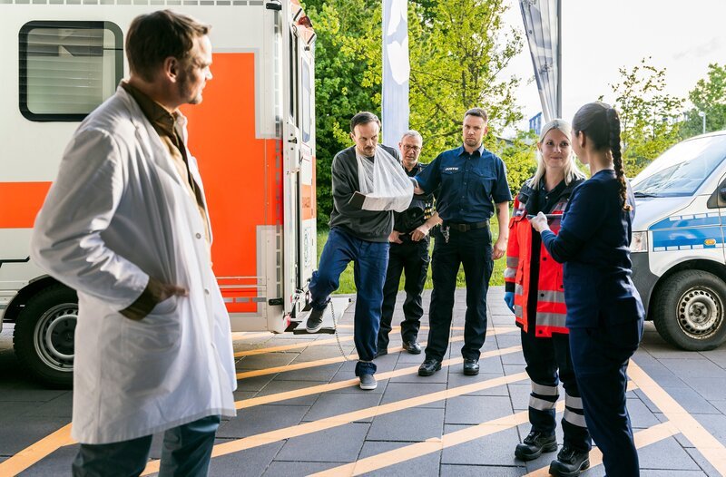 Dr. Marc Lindner (Christian Beermann) und Assistenzärztin Sofia Galura (Olivia Papoli-Barawati) nehmen Nils Staaken (Robert Besta) in der Notaufnahme auf. Dieser ist in Begleitung von Justizvollzugsbeamten (v.l.n.r.: Christian Beermann, Robert Besta, Komparsen und Komparsin). – Bild: ARD/​Michael Kremer