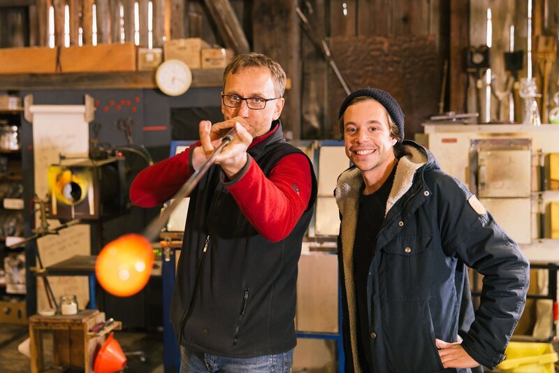 Glasbläser Stefan (links) zeigt Tobi in seiner Glasmanufaktur, wie Glas hergestellt wird. Ob ihm das bei der Lösung des CheXperiments hilft? – Bild: BR/​megaherz gmbh/​Hans-Florian Hopfner