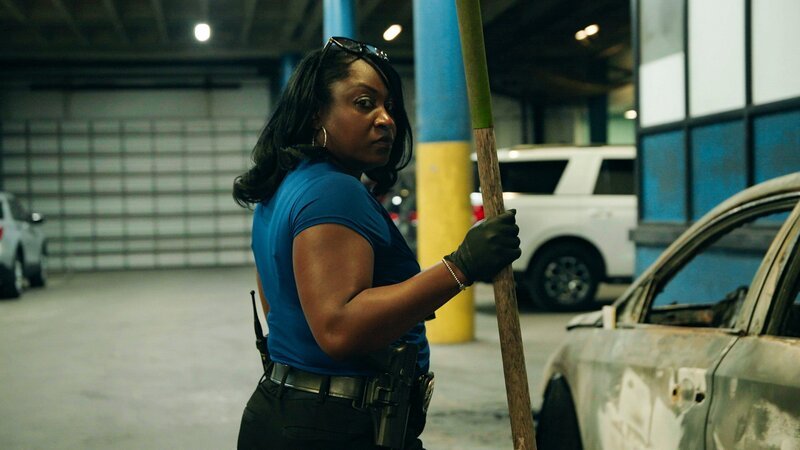 Eine Frau in einem blauen Shirt und Handschuhen hält einen langen Stab, während sie neben einem ausgebrannten Fahrzeug in einer Garage steht. – Bild: Crime + Investigation /​ A+E Global Media