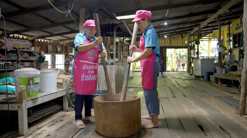Ein hundert Jahre alter Holz-Bottich und Baseballschläger-artige Stößel: Sunanta (links) und ihre Köchin Fern zerstampfen die kleinen Shrimps zu einer cremigen Würzpaste, die hier „Kapi“ heißt. – Bild: NDR/​Julian Ringer