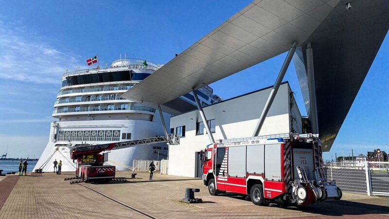 A firetruck at the cruise ship terminal Warnemünde. The turntable fire escape is extended to a window of the terminal. Behind the terminal lies a cruise ship. – Bild: Warner Bros. Discovery, Inc. or its subsidiaries and affiliates