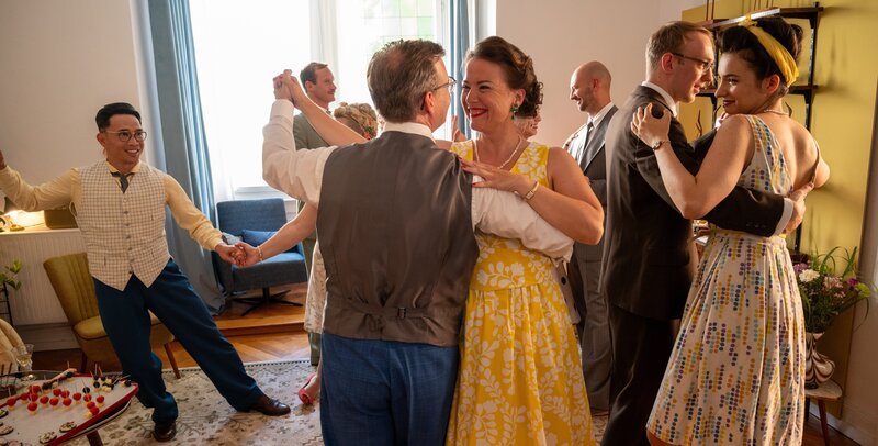 Während das Ehepaar Ina (Josephine Ehlert, 4.v.r.) und Georg Trapp (Dominik Klingberg, 4.v.l.) eine ausgelassene 50er-Jahre Party feiert, kommt es in dem Mehrfamilienhaus zu einer grausamen Tat: Am Fuß der Kellertreppe wird eine Frau tot aufgefunden – sie wurde gestoßen. Doch weder die Trapps, noch die Gäste (Komparsen) wollen etwas mitbekommen haben. – Bild: MARKUS_FENCHEL /​ ZDF und MARKUS_FENCHEL