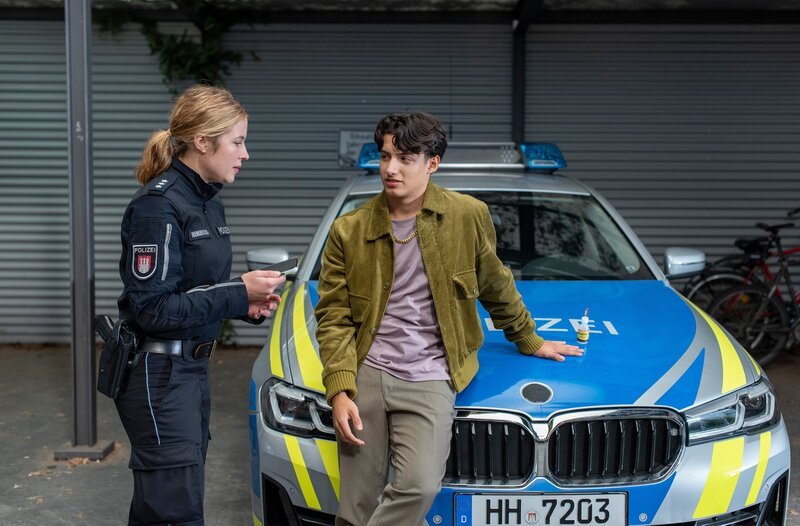 Keine Fotos mit Polizisten: Bente Hinrichs (Sinha Melina Gierke, l.) muss zu erzieherischen Maßnahmen greifen, um den liebestollen Mehmed (Giovanni Francesco, r.), der sich an einem Einsatzwagen festgeklebt hat, zur Besinnung zu bringen. – Bild: ARD/​Thorsten Jander