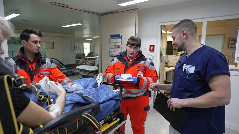 Lübeck: a pixelated female patient has just been brought to the A&E. Nurse Jacob is smilling and seems to be making the handover with two young paramedics, who are both wearing orange uniforms. They are at the A&E entrance. Behind them we see the staff room with a glass window and in the background several empty hospital gurneys. The female paramedic seems to be reading a note in her gloved hands. – Bild: Warner Bros. Discovery, Inc. or its subsidiaries and affiliates. All rights reserved. /​ Production Company Staff