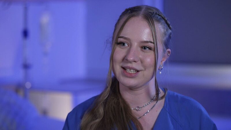 Kaufbeuren: close shot of young nurse Laura-Sophie, who is smiling while giving an interview on the interview set. The background is illuminated in shades of blue. She is wearing earrings and two necklaces. – Bild: Warner Bros. Discovery, Inc. or its subsidiaries and affiliates. All rights reserved. /​ Production Company Staff