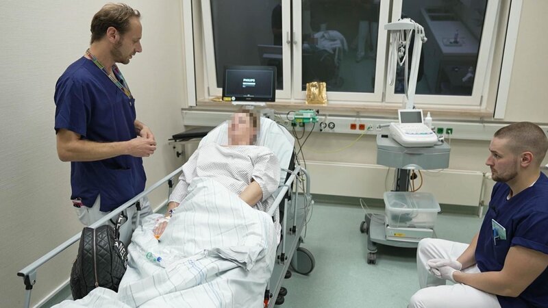 Lübeck: A female pixelated patient is laying on a bed, already wearing a hospital gown and seemingly having a conversation with nurse Florian. Nurse Jacob is listening to them while sitting on a nearby stool. Behind the bed we see the filming crew partially reflected on the big windows. A monitor and ultrasound device are also in the room. – Bild: Warner Bros. Discovery, Inc. or its subsidiaries and affiliates. All rights reserved. /​ Production Company Staff