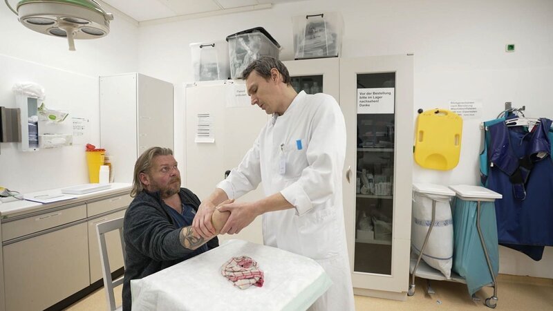 Lübeck: Physician Pavlo is checking on the tattood patient’s wrist. Pavlo is standing, the patient is looking up at him sitting at a small table. Behind them we see several white cupboards, to the right trash containers and protective x-ray coats – Bild: Warner Bros. Discovery, Inc. /​ Production Company Staff