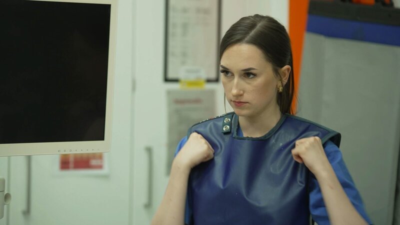 Riesa: close shot of nurse Charlotte while fixing her blue lead apron shortly before the operation begins. To her right there is a big turned off screen. Behind her lead curtins and something orange on the wall. – Bild: Warner Bros. Discovery, Inc. or its subsidiaries and affiliates. All rights reserved. /​ Production Company Staff