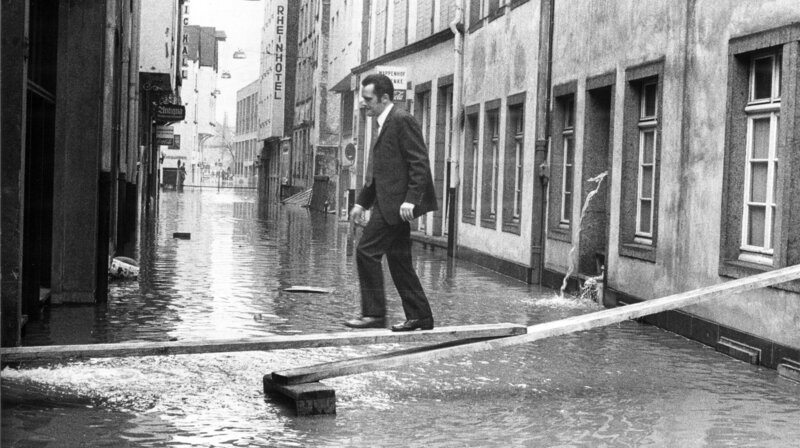 Hochwasser in der Kölner Altstadt am 25. Februar 1970: Ein Mann geht auf einem Steg aus Holzbohlen über eine der überschwemmten Straßen im Martinsviertel der Kölner Altstadt. Das Hochwasser erreichte an diesem Tag einen Pegelstand von 8,87 Metern. – Bild: WDR/​dpa