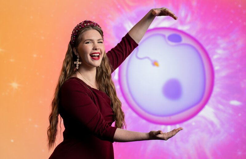 Host Melina Hoischen umarmt die auf dem Hintergrund gezeigte Eizelle. – Bild: Janne Ebel /​ DRIVE beta GmbH