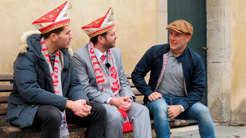 Oli trifft auf zwei Vertreter des Karnevalsvereins KG Alt Köln, die alte Kostüme mitbringen. – Bild: WDR/​Encanto Film- und Fernsehproduktions GmbH