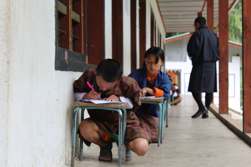 Tshering brütet über dem Mathetest, während ihr Lehrer kontrolliert. – Bild: SWR/​Thinley Namgay /​ KiKA