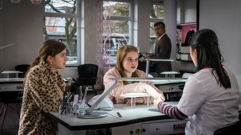 Lou (Luna Winter, li.) und Pippa (Elyza Silber, 2.v.li.) versuchen, mit Linhs Freundin Mai (My Dam, re.) Kontakt aufzunehmen, doch Nagelstudiochef Than (Lam Vissay) hat sie im Blick. – Bild: NDR/​Boris Laewen/​KiKA