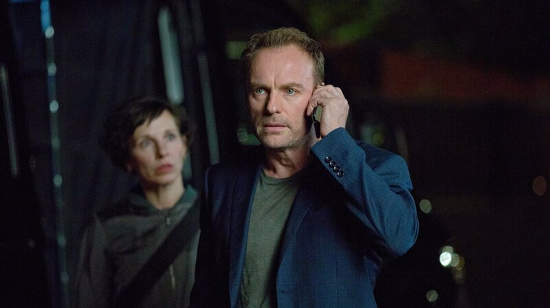 Ein belebter Platz mitten in der Stadt, 12 Uhr mittags. Gerade winkt die Studentin Mina Jiang (Yun Huang) noch ihrer Kommilitonin Luise (Paula Kroh) von weitem zu, als sie plötzlich tot zusammenbricht. Ein Schuss in den Hinterkopf führt die Kommissare Rubin (Meret Becker) und Karow (Mark Waschke) in die historische Mitte Berlins. – Karow (Mark Waschke) und Rubin (Meret Becker) versuchen das Geschehen in der Villa des Colloquiums zu belauschen. – Bild: rbb/​die film gmbh/​Volker Roloff