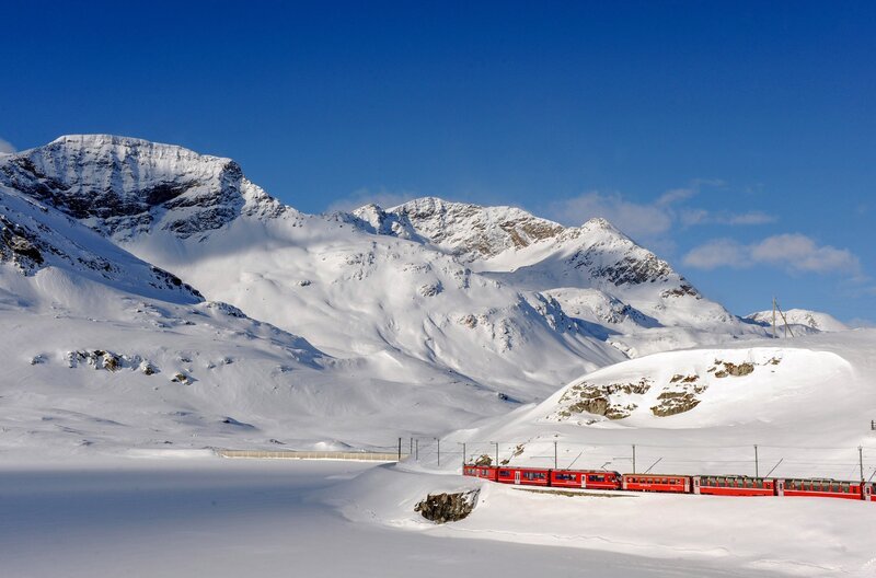 Der Bernina Express auf dem Berninapass am Lago Bianco: Der Zug schlängelt sich durch tiefe Täler, wilde Schluchten und über das ewige Eis der Bündner Alpen. – Bild: Rhätische Bahn /​ Tibert Keller