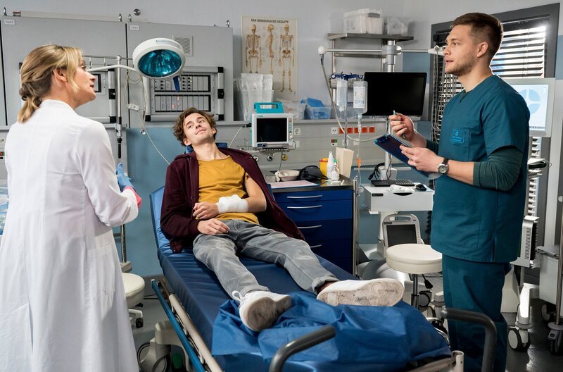 Lars Wexler (Leonard Fuchs, M.) wird nach einem Unfall in die Notaufnahme eingeliefert. Dr. Ruhland (Gunda Ebert, l.) ist als Belegärztin am JTK und übernimmt gemeinsam mit Mikko (Luan Gummich, r.) die Untersuchung. – Bild: ARD/​Jens Ulrich Koch